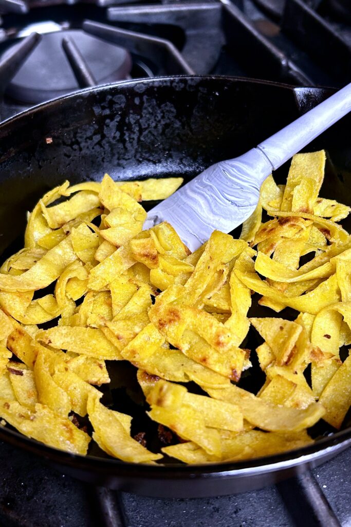 Tortilla cut into strips and gently tossed in lightly oiled black skillet. A small white spatula is towards the back of the pan used to stir the strips.